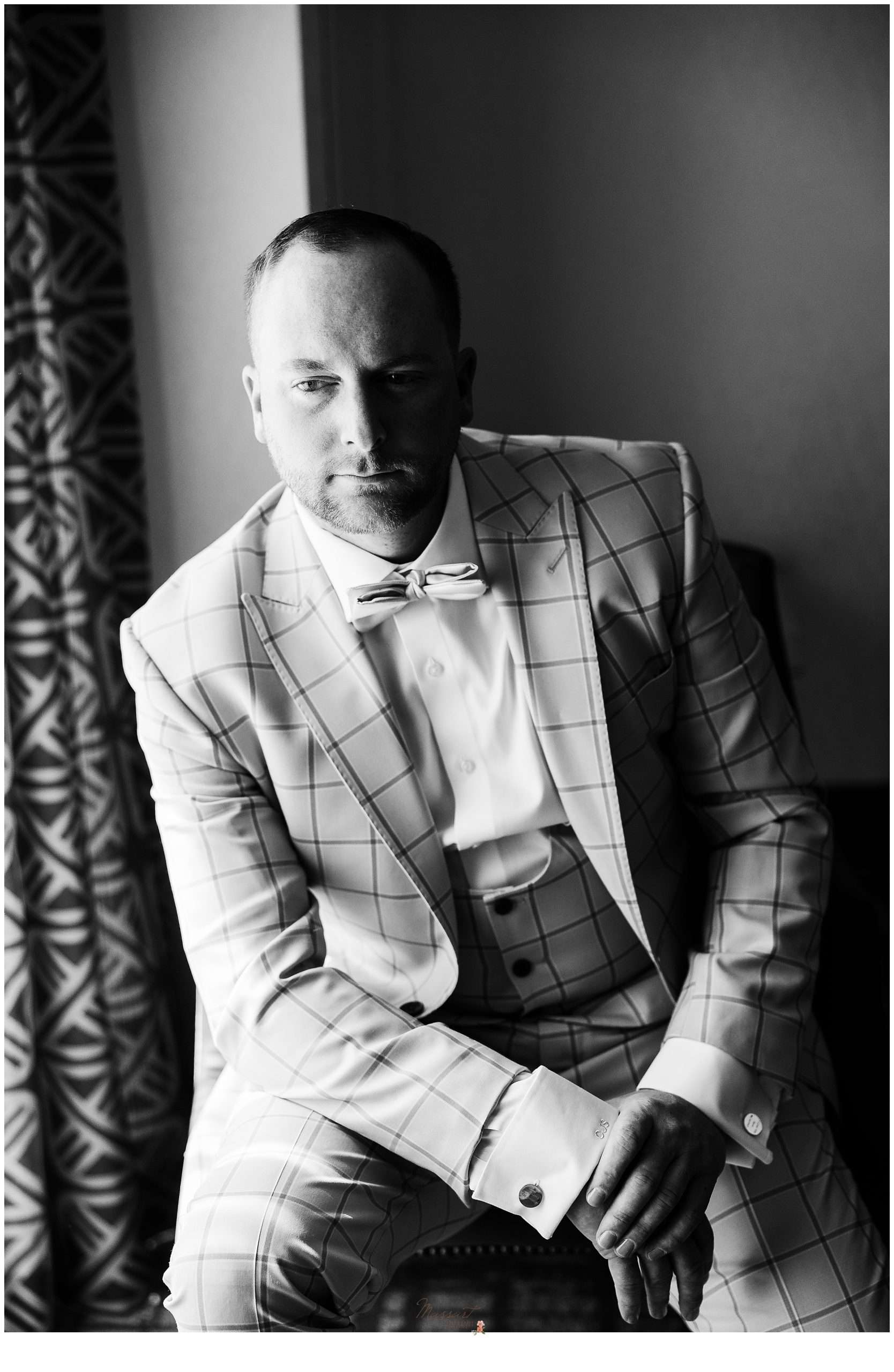 groom in tweed suit leans on knee sitting facing window in Rhode Island hotel room