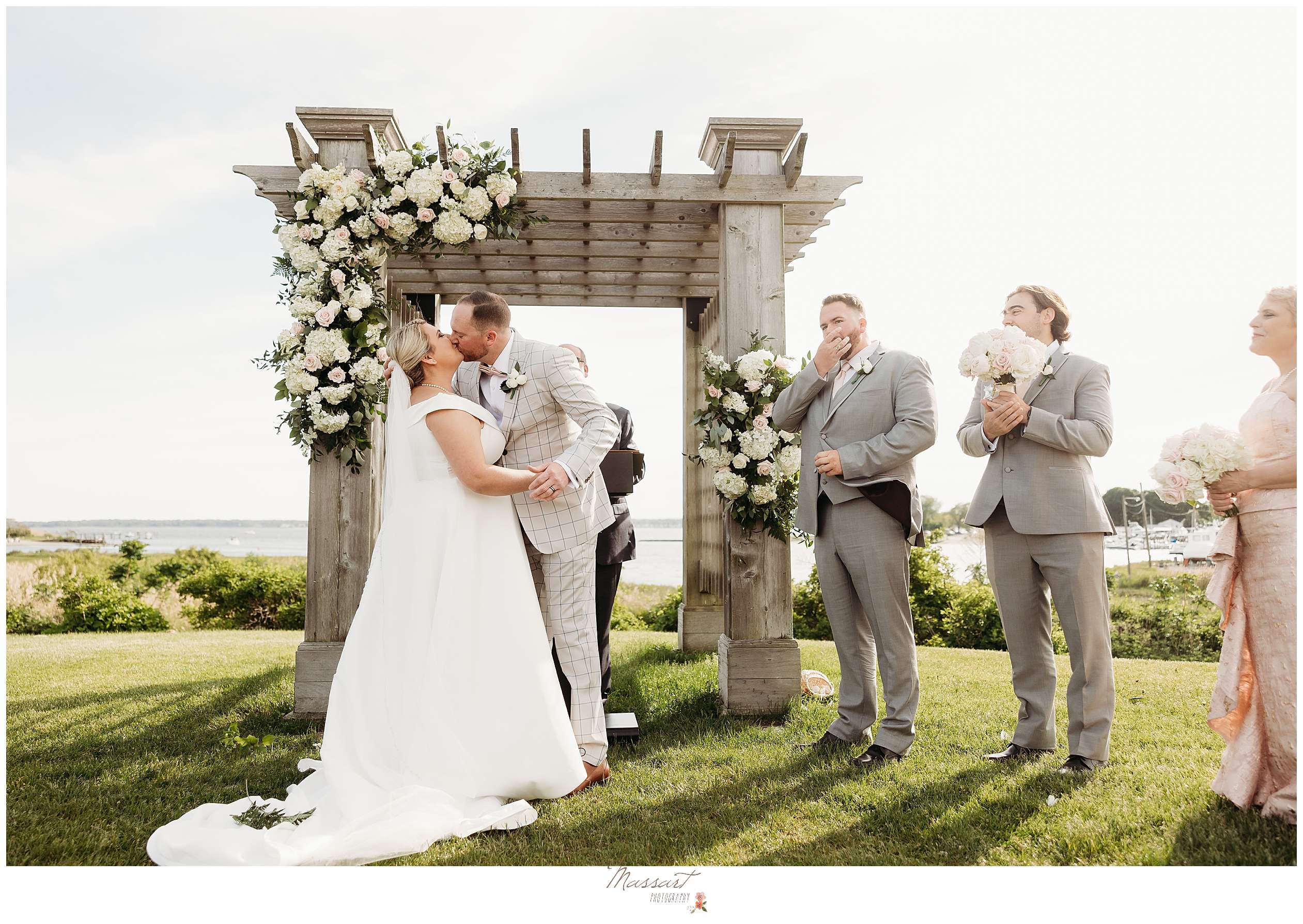bride and groom kiss by wooden arbor with white floral displays in front of waterfront at Harbor Lights
